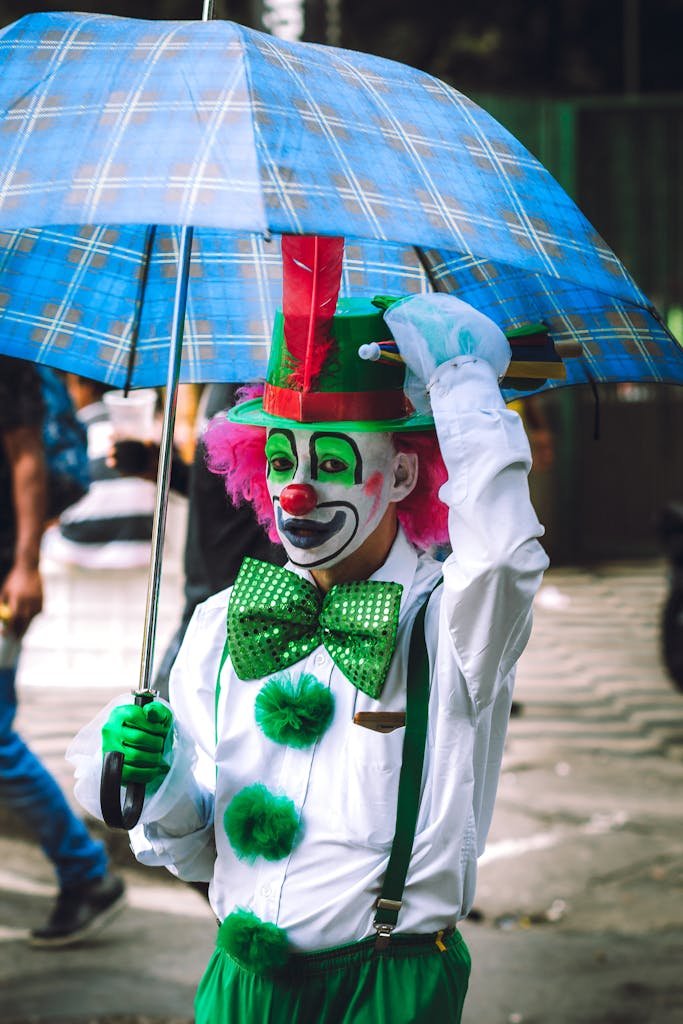 Anonymous male clown in funny costume and pink wig walking on street with umbrella and looking at camera