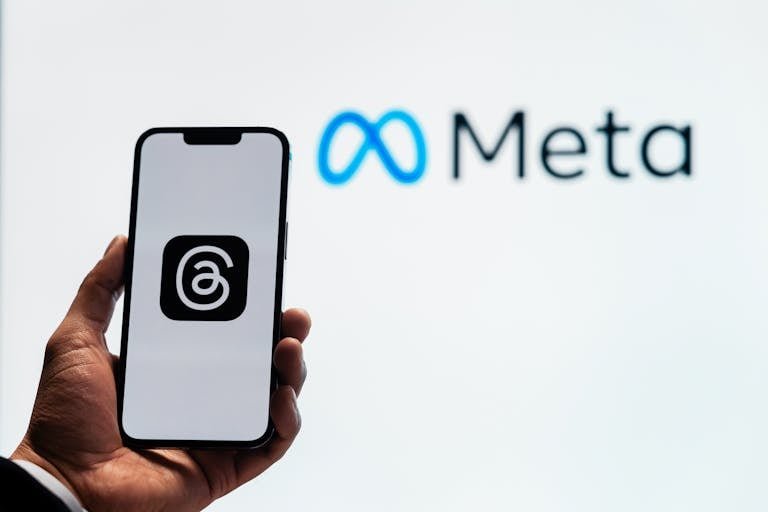 A hand holding a smartphone showing the Threads app with Meta logo in the background.